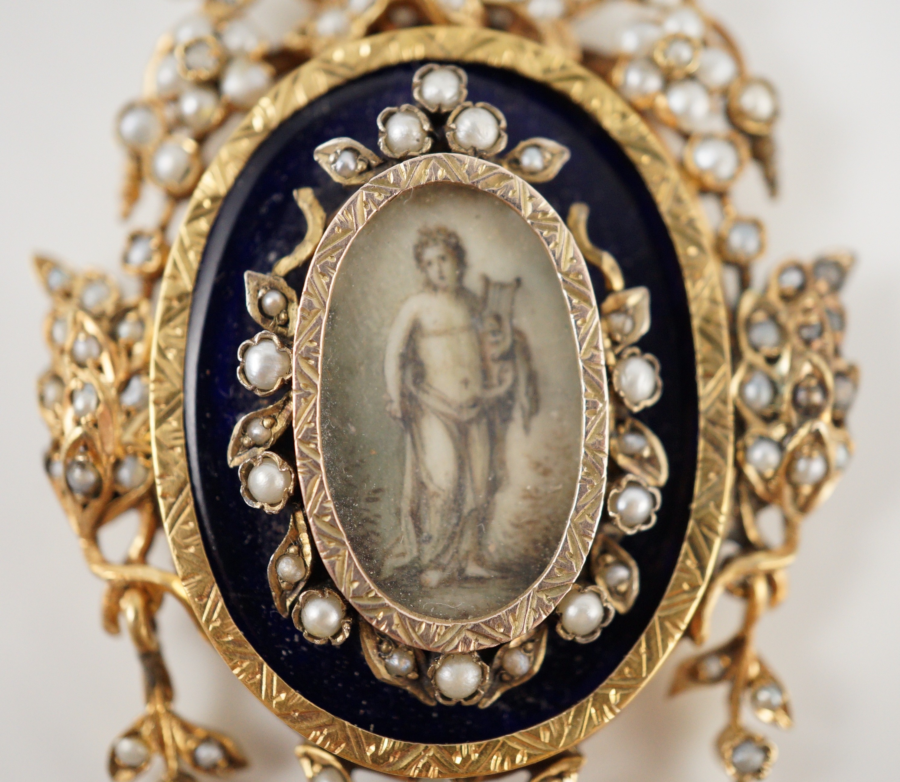 A Regency parcel gilt silver seed pearl and enamel set oval pendant, with inset ivory panel of a semi-clad figure with a lyre